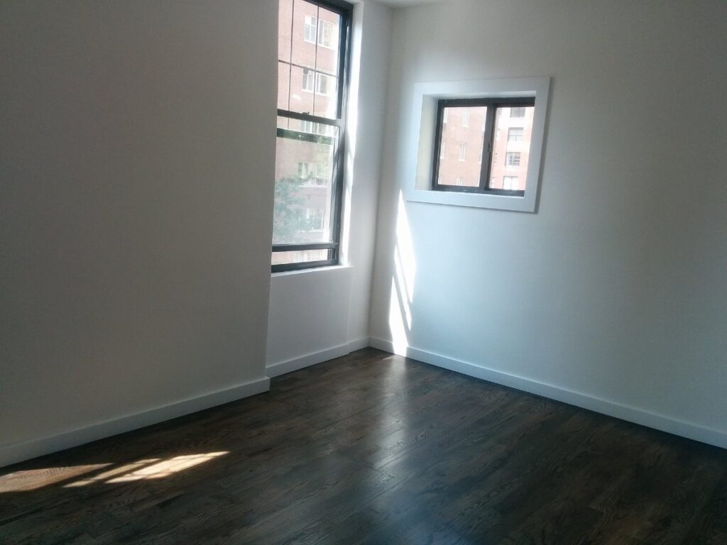 An empty, unfurnished apartment room with bare wooden flooring, white walls and daylight from two corner windows